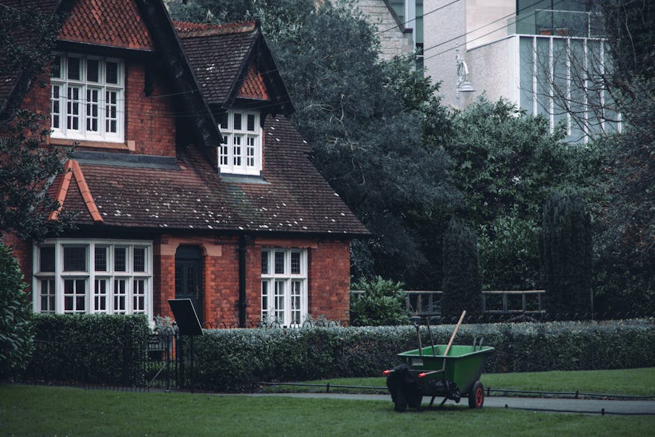A neatly maintained front garden of a traditional red brick house featuring a lush green lawn bordered by a low hedge. The garden includes a paved pathway alongside the hedge, with a green wheelbarrow