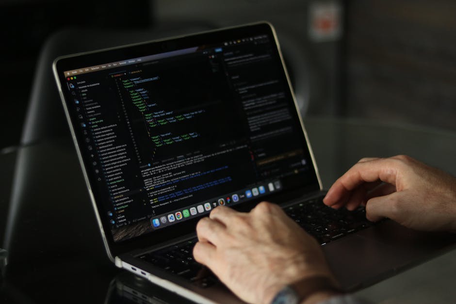A close-up view of a person's hands using a laptop on a dark, smooth surface. The laptop screen displays a code editor with lines of colorful code in various shades of green, blue, and white on a dark