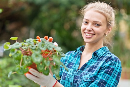 A young woman with light blonde hair tied back, smiling while holding a red container of vibrant green and reddish foliage plants, in an outdoor garden setting with blurred background of trees and shrubs, suggesting active gardening or planting activity. She is dressed casually in a blue checked shirt and wearing gardening gloves, indicating involvement in garden maintenance or planting at a residential garden or landscaped outdoor space near Abbey Wood. The natural lighting highlights the lush greenery and creates a welcoming, professional environment associated with gardening services offered by Lawn Mowing Abbey Wood, emphasizing outdoor plant care and garden enhancement in a suburban setting.